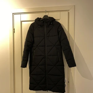 Svart lång dunjacka från Zero - Snygg och varm svart lång dunjacka från Zero. Jackan har en dragkedja framtill och långa ärmar. Perfekt för kalla dagar med sin quiltade design som ger extra värme och stil.