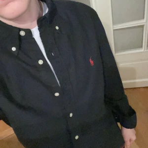Svart skjorta från Ralph Lauren - Snygg svart skjorta från Ralph Lauren med klassisk krage och knappar framtill. Skjortan har en broderad röd logotyp på bröstet och är långärmad. Perfekt för en stilren look.
