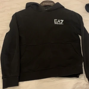 Svart hoodie från EA7 Emporio Armani - Säljer en stilren svart hoodie från EA7 Emporio Armani. Tröjan har en klassisk design med en känguruficka framtill och en logotyp på bröstet. Perfekt för en avslappnad look. Pris kan diskuteras via köp!