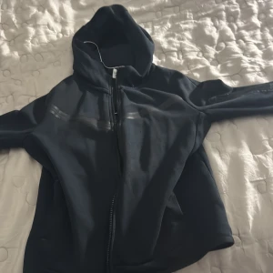 Tech fleese svart  - Säljer en stilren svart vindjacka med huva och dragkedja framtill. Perfekt för blåsiga dagar och har en sportig look. Jackan har långa ärmar och är tillverkad i ett syntetmaterial som skyddar mot väder och vind.