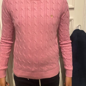 Rosa kabelstickad tröja från Morris - Säljer en snygg rosa kabelstickad tröja från Morris Lady i 100% Pima Cotton. Tröjan har långa ärmar och ett diskret broderat emblem på bröstet. Perfekt för en stilren look!