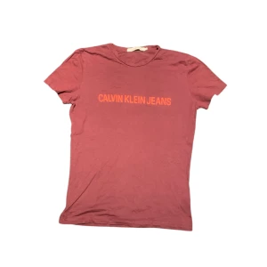 Röd t-shirt från Calvin Klein Jeans - Snygg röd t-shirt från Calvin Klein Jeans med tryck i orange på framsidan. Perfekt för en avslappnad stil. Tillverkad i mjukt material för extra komfort.