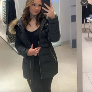 HOLLIES JACKA - Snygg svart dunjacka med pälskrage och dragkedja. Perfekt för kalla dagar med sin långa design och varma material. Jackan har en figurnära passform och praktiska fickor.