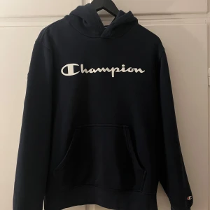 Marinblå hoodie från Champion - Säljer en marinblå hoodie från Champion med deras ikoniska logga i vitt på bröstet. Tröjan har en klassisk känguruficka och en bekväm huva. Perfekt för en avslappnad stil. Ett hål tyvärr i ena ärmen därför pris 