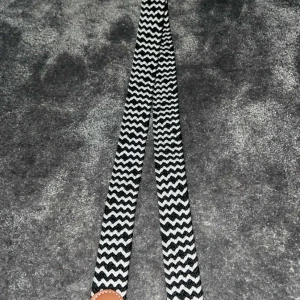 Svartvitt bälte med zigzag-mönster - Snyggt svartvitt bälte med ett coolt zigzag-mönster. Perfekt för att ge din outfit en unik touch. Bältet har en brun detalj vid spännet som ger en fin kontrast. Passar till många olika stilar och tillfällen. FRÅN ONEWEAR