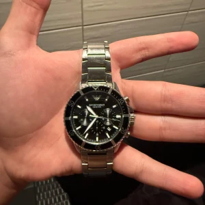  Klocka Emporio Armani (Dive) - Snygg klocka från Emporio Armani med svart urtavla och silverfärgad länkarmband. Klockan har tydliga markeringar och en stilren design som passar perfekt för den modemedvetna. Perfekt accessoar för att lyfta din stil.