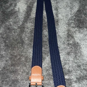 Blått flätat bälte  - Snyggt blått flätat bälte med bruna läderdetaljer. Perfekt för att ge en stilren touch till din outfit. Bältet har en klassisk design med en robust spänne. FRÅN ONEWEAR 