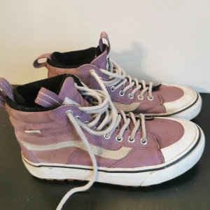 Lila sneakers från Vans - Snygga lila sneakers från Vans med högre skaft och klassisk vit sula. Skorna har snörning och en vit sidostrimma som ger en ikonisk look. Perfekta för en avslappnad stil.
