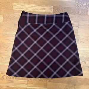 Rutig kjol med dragkedja - Snygg rutig kjol i mörka nyanser med dragkedja på sidan. Perfekt för en stilren look. Kjolen har ett klassiskt rutmönster i svart och lila.