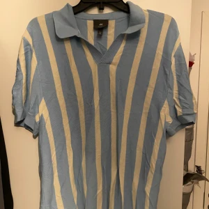Blå och vit randig pikétröja från H&M - Säljer en stilren blå och vit randig pikétröja från H&M. Tröjan har korta ärmar och en klassisk krage. Perfekt för en avslappnad stil. Hör av dig vid intresse!