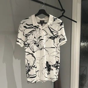 Vit och svart pikétröja från H&M - Säljer en snygg vit pikétröja från H&M med svart blommönster. Tröjan har en klassisk krage och korta ärmar, perfekt för en stilren look. Passar både till vardags och festliga tillfällen. 🖤