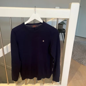 Mörkblå tröja från Morris - Säljer en stilren mörkblå tröja från Morris i 100% merinoull. Perfekt för både höst och vinter med sin mjuka och varma känsla. Tröjan har en liten broderad logga på bröstet och ribbade kanter vid ärmslut och nederkant. Passar både till vardags och lite finare tillfällen.
