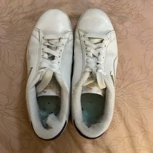 Vita sneakers från Puma - Säljer ett par vita Puma sneakers i skinn med klassisk design och snörning. De har en diskret guldfärgad logga på sidan och är i bra skick. 