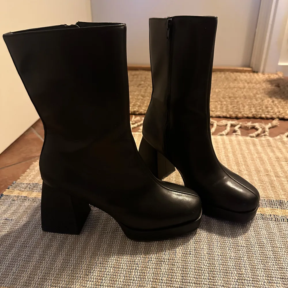 Snygga svarta boots i skinn med en rejäl klack och dragkedja på sidan. Perfekta för höst och vinter, dessa boots ger en stilren look och är superbekväma att gå i. De har en modern design med en fyrkantig tå som ger extra edge till din outfit.. Skor.