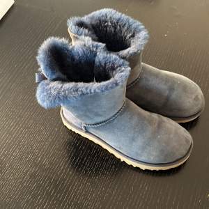 Säljer ett par mysiga blå UGG boots i mycket bra skick. De har en fluffig insida och en söt rosett på baksidan. Perfekta för kalla dagar och ger en stilren look. Sulan är robust och ger bra grepp. Passar perfekt för höst och vinter!