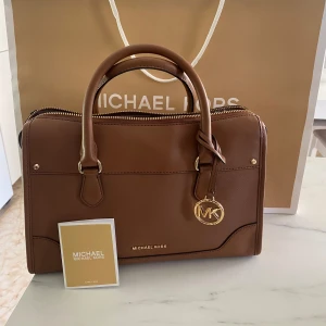 Brun handväska från Michael Kors - Snygg brun handväska från Michael Kors i skinn. Väskan har en elegant design med dragkedja och ett dekorativt MK-hänge. Perfekt för både vardag och fest!