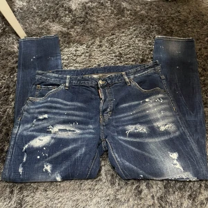 Blå jeans från DSQUARED2 - Säljer ett par blå DSQUARED2 jeans i storlek 54. De har en cool distressed look med slitningar och färgstänk. Perfekta för en avslappnad och trendig stil. Passar bra till både vardag och fest!