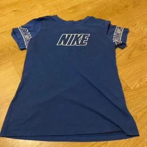 Blå Nike T-shirt - Säljer en blå Nike T-shirt i Dri-FIT material, perfekt för träning. Den har korta ärmar med ett coolt mönster och Nike-loggan tryckt på bröstet. Skön och andas bra, idealisk för aktiva dagar. Passar både till vardags och sportiga aktiviteter.