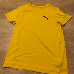 Gul t-shirt från Puma - Säljer en snygg gul t-shirt från Puma i bomull. Den har en klassisk passform med korta ärmar och en svart Puma-logga på bröstet. Perfekt för sommardagar eller träning. 