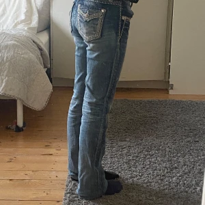 Miss Me med detaljer - Säljer ett par snygga blå jeans från Miss Me med coola broderade detaljer och silverknappar. Bootcut, storlek 26. Jeansen har en unik design på bakfickorna som verkligen sticker ut. Perfekt för dig som vill ha något extra i garderoben! 💙