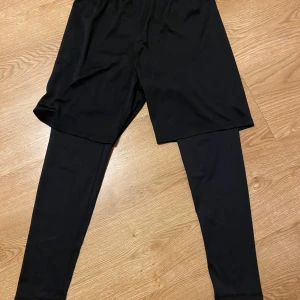 Svarta sportshorts med tights från H&M - Säljer ett par svarta sportshorts med inbyggda tights från H&M. Perfekta för träning eller löpning. De är i bra skick och har en bekväm passform. Passar både för inomhus- och utomhusaktiviteter.