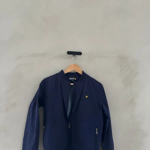 Mörkblå bomberjacka från Lyle & Scott - Säljer en snygg mörkblå bomberjacka från Lyle & Scott. Den har en dragkedja framtill och långa ärmar. Perfekt för höst och vår! Jackan har en liten gul logga på bröstet som ger en stilren touch. Passar både till vardags och fest.