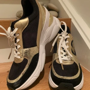 Svart och guld sneakers från Michael Kors med kilklack - Snygga sneakers i svart och guld från Michael Kors. De har en kilklack vit sula och är dekorerade med gulddetaljer och ett stort MK-logotyp på sidan. Skorna har vita snören och en glittrig finish som gör dem perfekta för både vardag och fest. De är i mycket bra skick och redo att bäras med stil! Storlek 39