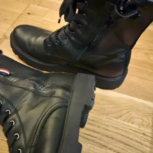 Snygga svarta boots i skinn från Tommy Hilfiger. De har snörning och dragkedja på sidan för enkel på- och avtagning. Perfekta för höst och vinter med en robust sula som ger bra grepp. Klassisk design med märkets logga på sidan och detaljer i rött, vitt och blått.