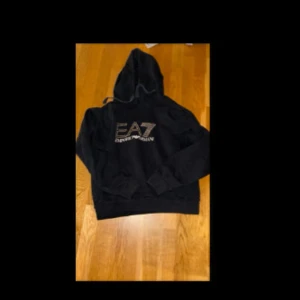 Svart hoodie från EA7 - Säljer en snygg svart hoodie från EA7 Emporio Armani. Den har en stor logga på bröstet och en klassisk känguruficka framtill. Perfekt för en avslappnad stil eller träning. Huvan har dragsko för justerbar passform.