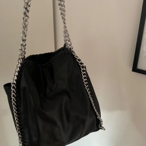 Svart axelväska med kedjedetaljer - Snygg och rymlig svart axelväska i skinn med coola kedjedetaljer. Har en liten söm som lossnat (bild 3) men går att sy fast så är det inget man tänker på! 