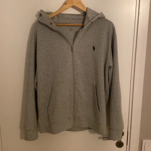 Grå hoodie från Ralph Lauren - Säljer en snygg och bekväm grå hoodie från Ralph Lauren. Den har en dragkedja framtill och en klassisk logga på bröstet. Perfekt för både vardag och chill. Passar bra till jeans eller joggers!