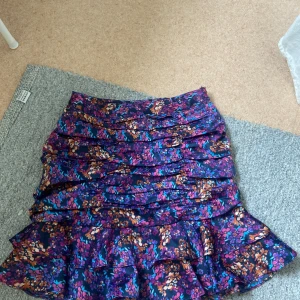 Blommig kjol från Vero Moda -  Vero Moda i storlek S. Kjolen har ett färgglatt mönster med lila, rosa och orange blommor på en mörkblå bakgrund. Den har volanger och en dragkedja i sidan. 