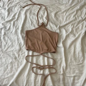 Beige halterneck topp - Säljer en snygg beige halterneck topp med knytband som går att vira runt midjan. Perfekt för sommaren och festliga tillfällen. Toppen är i ett mjukt och bekvämt material.