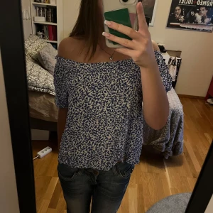 Blå blommig offshoulder-top - Säljer en superfin blå offshoulder-top med blommigt mönster. Den har korta ärmar och en luftig passform. Toppen är i ett mjukt och bekvämt material.