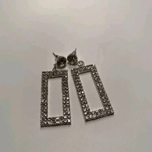 Glittriga rektangulära örhängen - Säljer ett par superglittriga rektangulära örhängen i silverfärg. De är perfekta för att lysa upp vilken outfit som helst och har en modern, stilren design. Örhängena är rektangulära och täckta med små gnistrande stenar