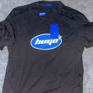 Svart t-shirt från Hugo - Säljer en svart t-shirt från Hugo i nytt skick. Den har en cool blå logga på bröstet och är perfekt för vardagsbruk. T-shirten är kortärmad och gjord i ett mjukt material som känns skönt mot huden. Passar perfekt till jeans eller shorts!