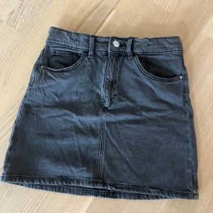 Svart/grå jeanskjol - Snygg svart jeanskjol i kort modell med klassisk ficksdesign. Perfekt för vardagsbruk. Kjolen har en knapp och dragkedja framtill för enkel påklädning. Passar bra till både t-shirt och blus! Den är i storlek 152