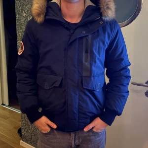 Mörkblå vinterjacka med pälsdetalj - Säljer en mörkblå vinterjacka med pälsdetalj på huvan. Jackan har en dragkedja och knappar framtill, samt en cool patch på ärmen. Pälsdetaljen går att sätta på och av.