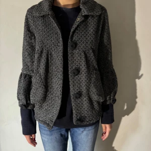 Grå glitter mönstrad jacka - Säljer en snygg grå jacka med ett unikt glittrigt mönster. Den har långa ärmar och stora knappar framtill. Perfekt för höst och vår, och ger en stilren look till vardagsoutfiten. Jackan är i bra skick och passar perfekt för vardagen och fest
