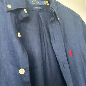 Blå linneskjorta från Ralph Lauren - Säljer en snygg blå linneskjorta från Ralph Lauren i storlek S. Skjortan har en klassisk krage och knappar framtill. Den är långärmad och har Ralph Laurens ikoniska röda logga broderad på bröstet. Perfekt för både vardag och fest! 💙skjortan är aldrig använd så är i jättefint skick 