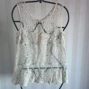 Vit spetslinne ny  - Säljer ett superfint vitt spetslinne med vackra detaljer. Det är ärmlöst och har en dragkedja i ryggen. Perfekt för sommardagar eller festliga tillfällen. Linnet har ett unikt mönster med både blommor och cirklar, vilket ger det en romantisk känsla. Passar perfekt till jeans eller kjol för en chic look! Ny