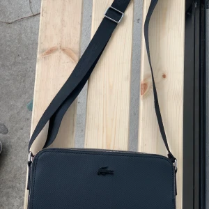 Svart axelväska från Lacoste - Snygg och stilren svart axelväska från Lacoste i mycket bra skick. Väskan har en justerbar axelrem och en praktisk dragkedja upptill. Perfekt för både vardag och fest! 🖤