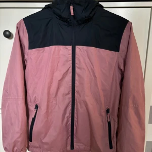 Rosa och marinblå vindjacka från McKinley - Säljer en snygg rosa och marinblå vindjacka från McKinley. Perfekt för blåsiga och regniga dagar med sin lätta och bekväma design. Jackan har en praktisk huva och dragkedja framtill. Den är i mycket bra skick och redo för nya äventyr!
