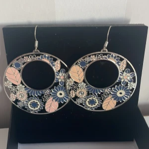Blommiga örhängen i silver och blått - Säljer ett par stora, runda örhängen i silver med ett vackert blommönster i blått och rosa. De har en öppen design med detaljerade blommor och blad som ger en bohemisk känsla. Perfekta för att piffa upp en outfit till fest eller vardag! 🌸