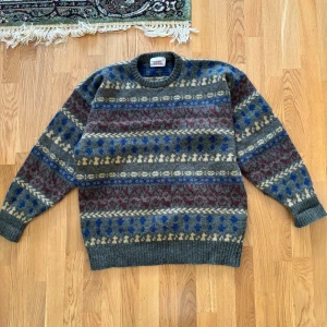 Mönstrad ulltröja i Shetland ull - Säljer en vintage mönstrad tröja från Commander i 100% Shetlandsull. Den har ett coolt mönster i grått, blått, rött och beige. Perfekt för kalla dagar med sin varma och sköna känsla. Tröjan har långa ärmar och ribbade kanter. Passar perfekt för höst och vinter!