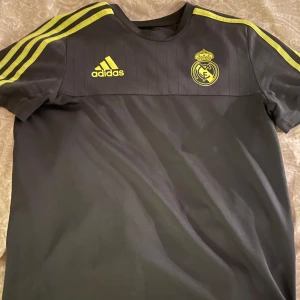 Grå Real Madrid träningströja från Adidas - Säljer en grå Real Madrid träningströja från Adidas i bra skick. Den har korta ärmar och gula detaljer, inklusive tre ränder på axlarna och klubbmärket på bröstet. Perfekt för fotbollsträning eller som en del av din supporteroutfit. Skön och stilren design!