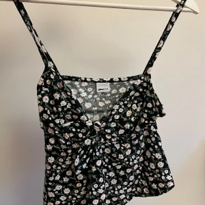 Blommig linnetopp från Gina Tricot - Söt blommig linnetopp från Gina Tricot i svart med vita och rosa blommor. Toppen har smala axelband och en knytdetalj framtill. Perfekt för sommardagar eller en avslappnad vårlook. Passar bra till jeans eller kjol 
