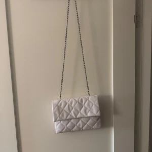 Vit quiltad axelväska - Säljer en superfin vit quiltad axelväska med kedjerem. Väskan har två fack och stängs med tryckknappar. Perfekt för både vardag och fest! 👜✨