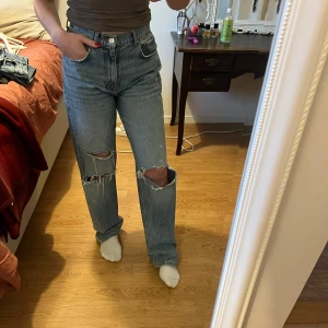 Blå jeans med slitningar - Säljer ett par snygga blå jeans med slitningar på knäna. De har en hög midja och en straight passform som ger en avslappnad look.