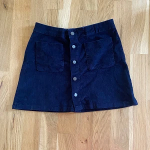 Svart manchesterkjol med knappar - Snygg svart manchesterkjol i kort modell med knappar framtill. Kjolen har två stora fickor och en skön passform som passar perfekt för både vardag och fest. Ett måste i garderoben för den som älskar klassiska plagg med en twist!